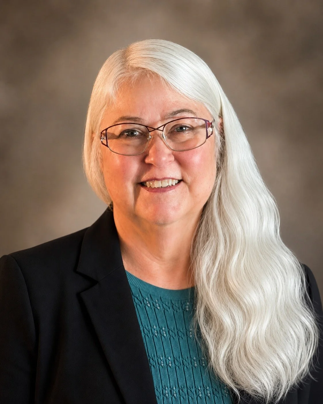 Portrait of Pam  Chandler, Bookkeeper with long white hair, glasses, and wearing a black blazer over a teal top, against a neutral background.