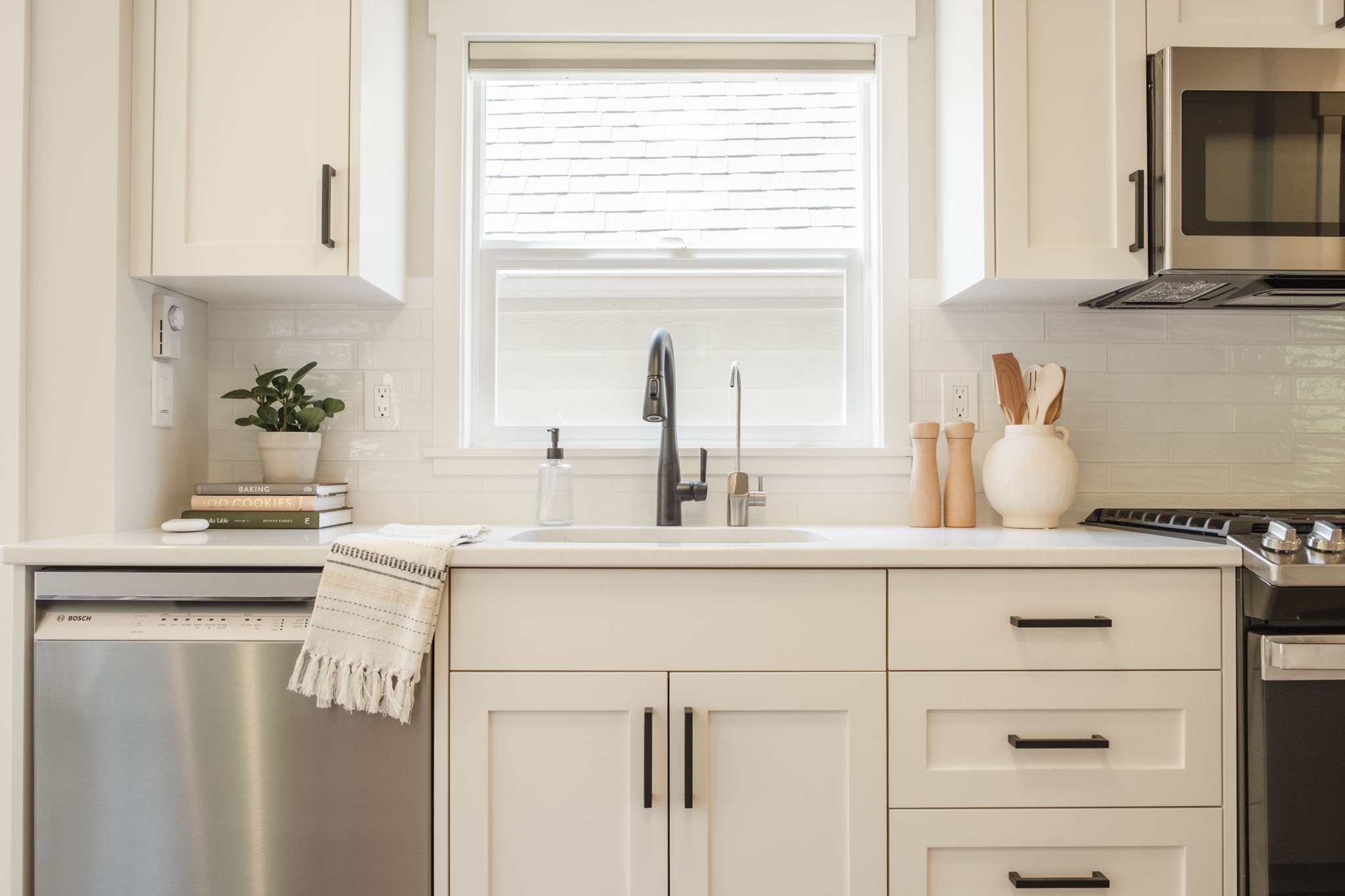A modern kitchen with white cabinets, a window above the sink, a plant, books, and kitchen utensils on the counter.