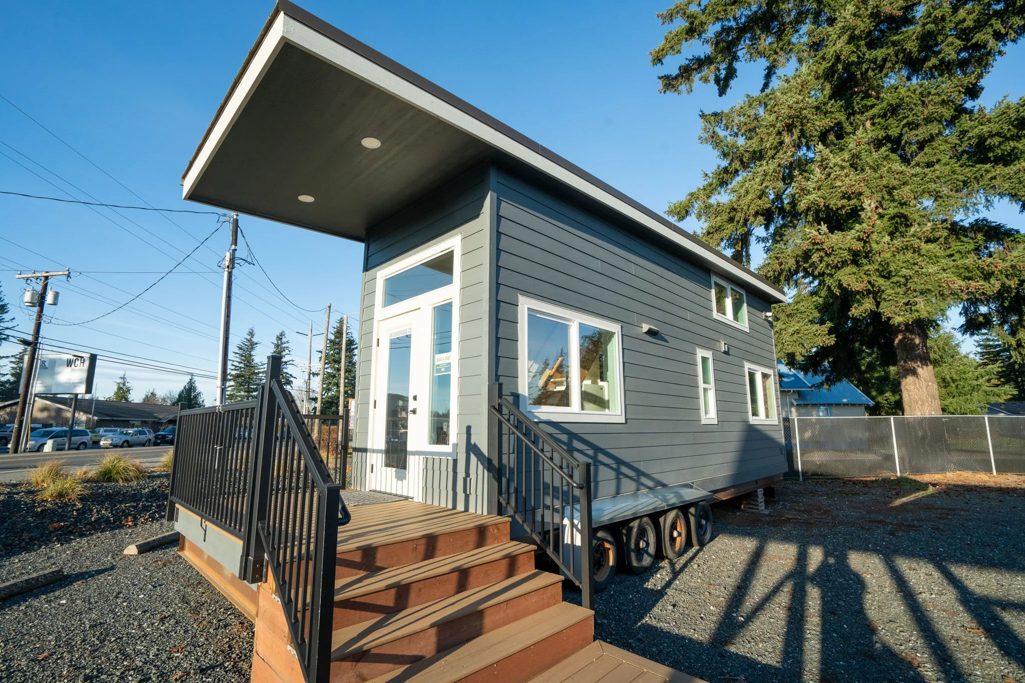 A tiny modern house on a trailer with a wooden staircase leading to the front door. The house has gray siding and multiple windows, with a large tree and power lines in the background.