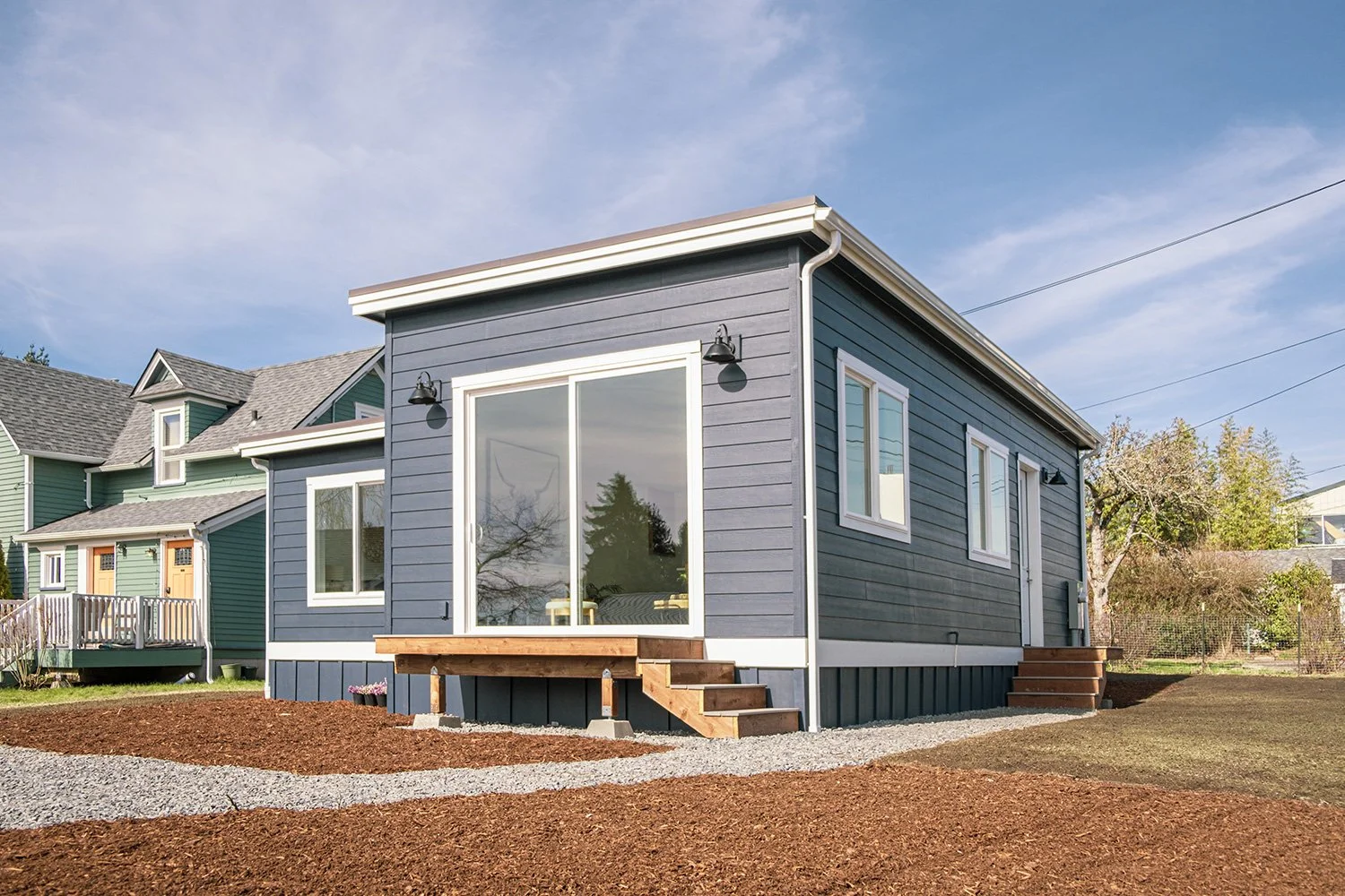 Blue house with white trim and sliding glass door, wooden stairs, surrounded by a landscaped yard with mulch and gravel, neighboring houses, trees, and power lines under a blue sky.