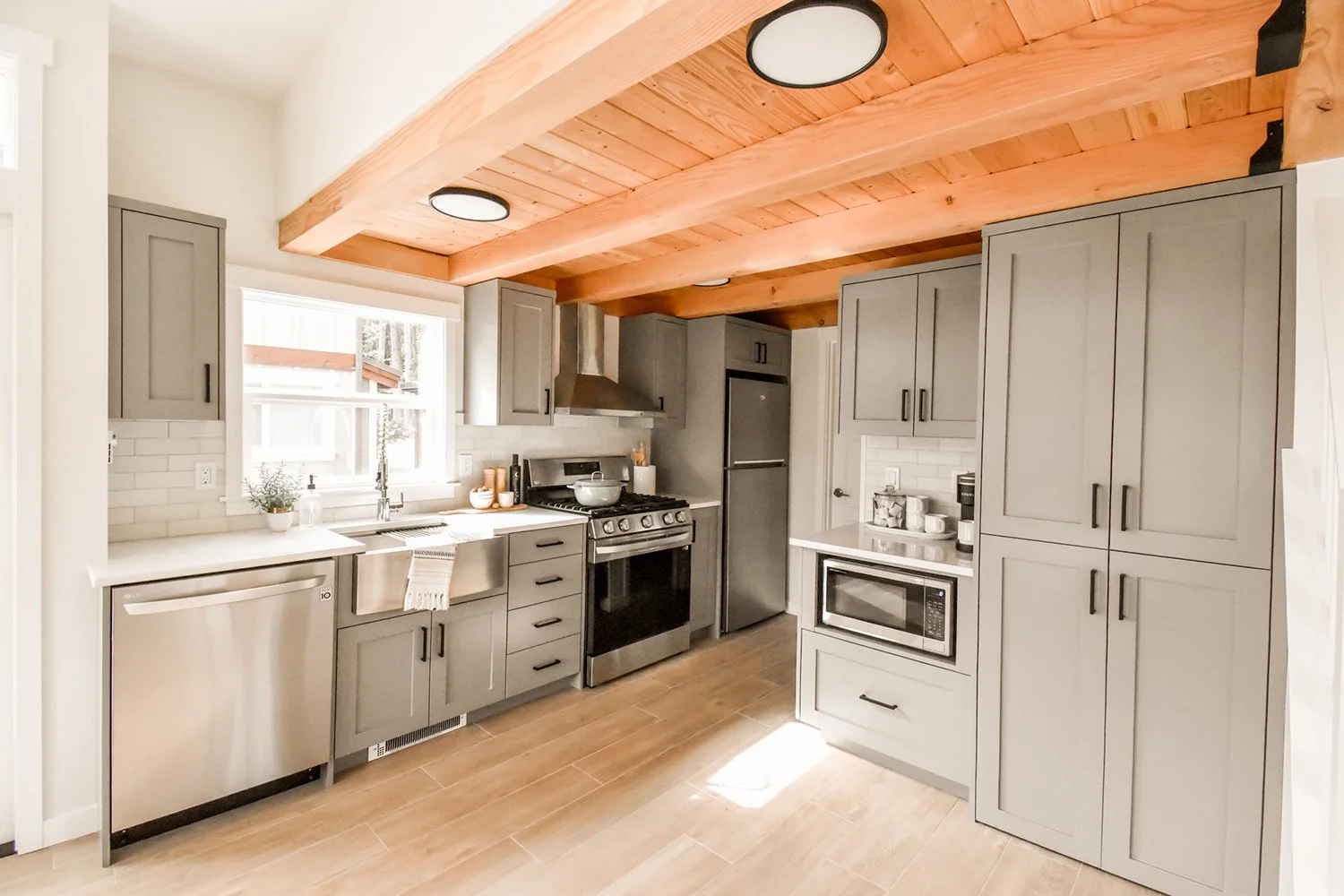 Modern kitchen with gray cabinets, stainless steel appliances, a window above the sink, and a wooden ceiling beam.