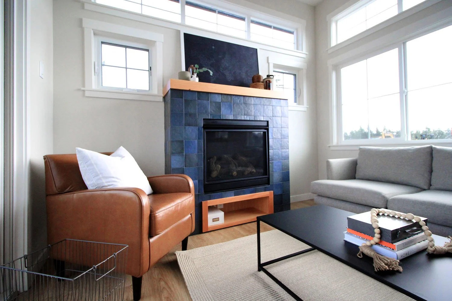 Living room with large windows, gray sofa, brown leather armchair with white pillow, black coffee table with books and decorative bead garland, and modern blue-tiled fireplace with black glass door and wooden mantel.