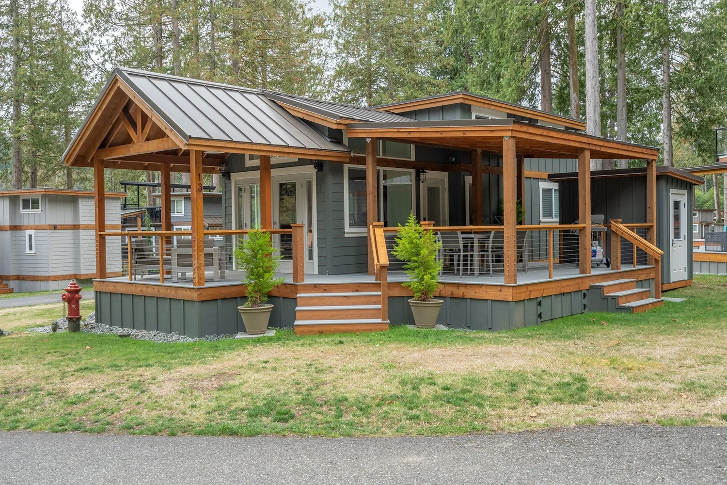 A modern house with a spacious wraparound deck, supported by wooden beams, real patio furniture, potted plants, large trees in the background, and a neighboring similar house.