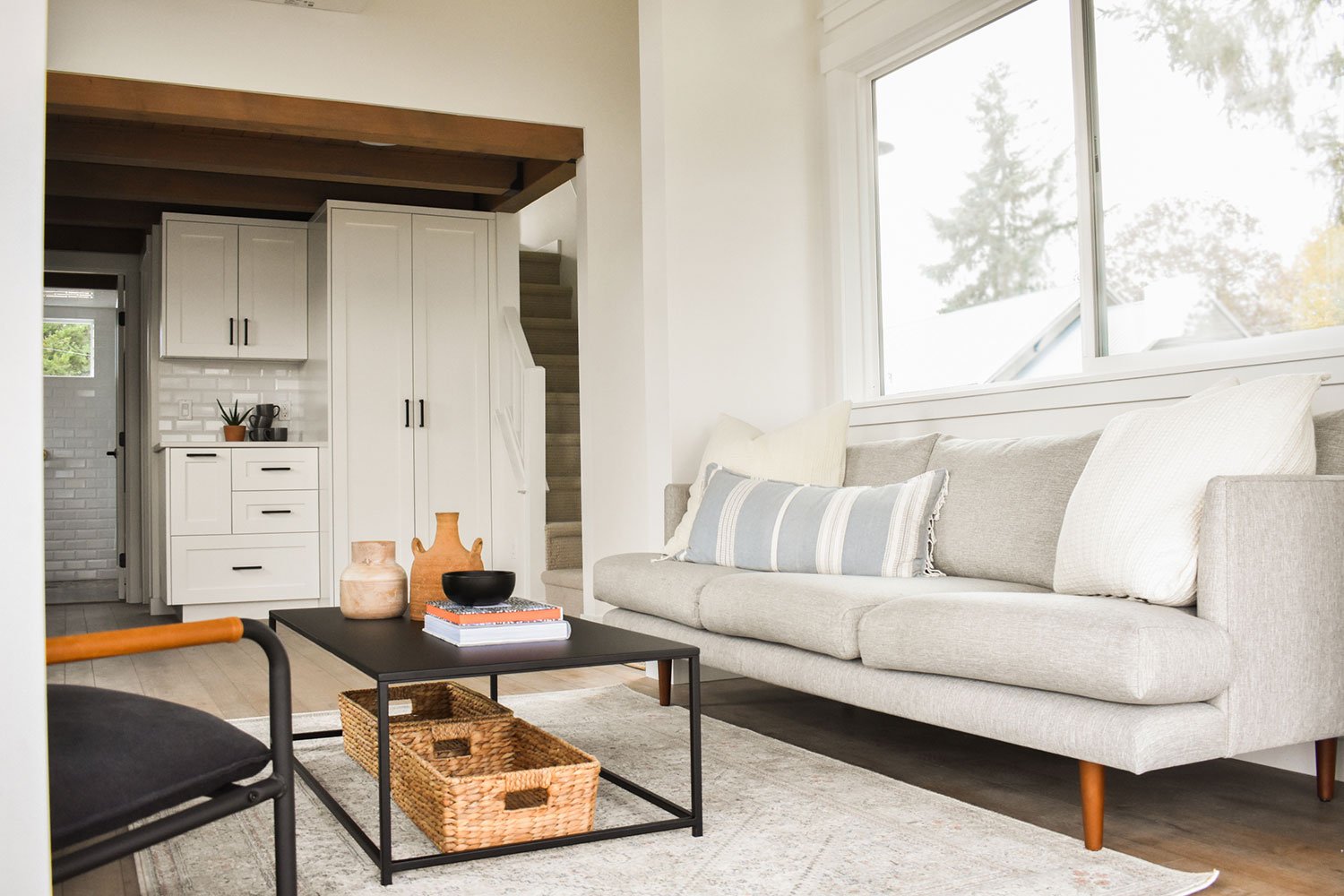Living room with a light gray sofa with white and striped pillows, a black coffee table with decorative vases and books, and a large window bringing in natural light. In the background, a staircase leading up to the second floor and a glimpse of a kitchen area with white cabinets.