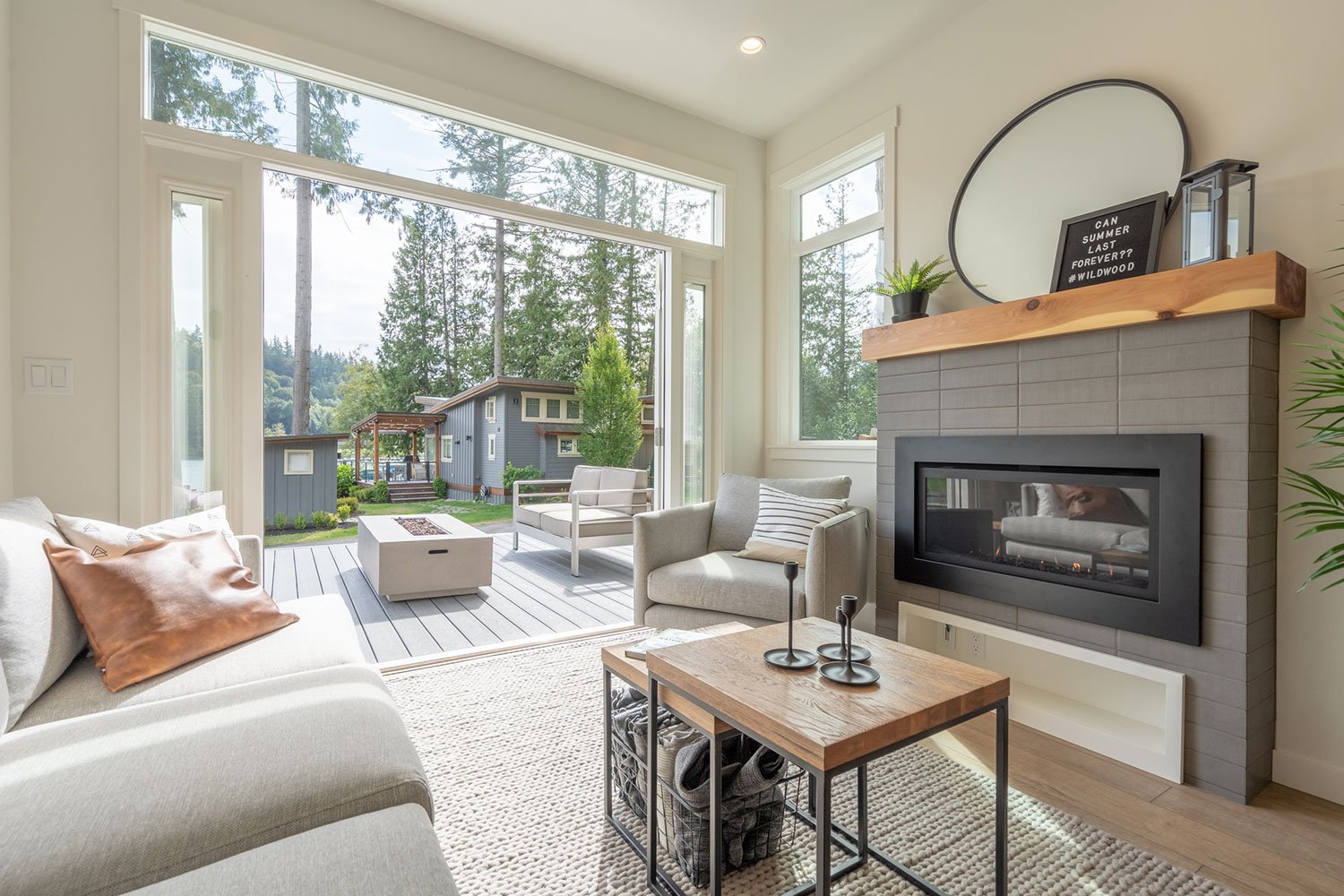 A living room with a fireplace, grey furniture, and a large sliding glass door leading to an outdoor deck. Outside, there are trees and a blue house.
