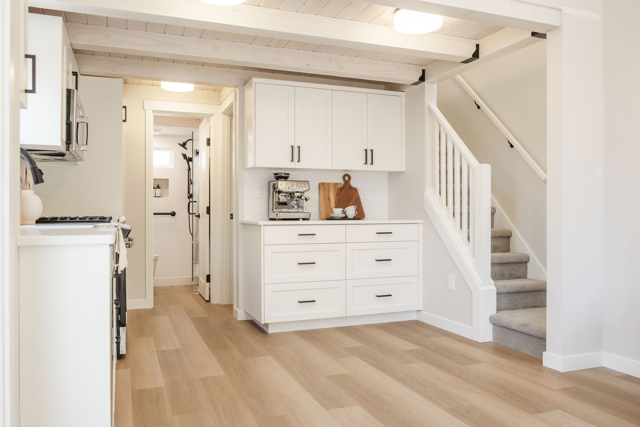 Interior view of a modern kitchen with white cabinets, a coffee machine on the counter, a wooden cutting board with cups, beige flooring, staircase with carpeted steps, and ceiling beams.