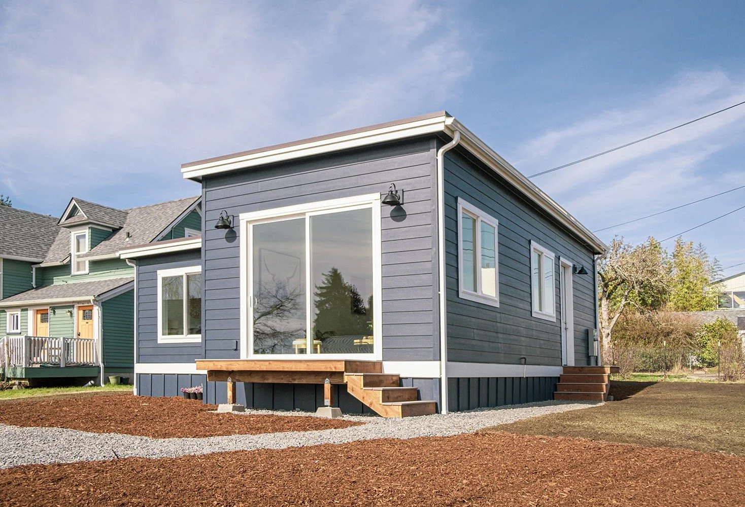 Front view of a newly built blue house with white trim, small yard with gravel walkway, and steps leading to front door and side door. Clear sky.