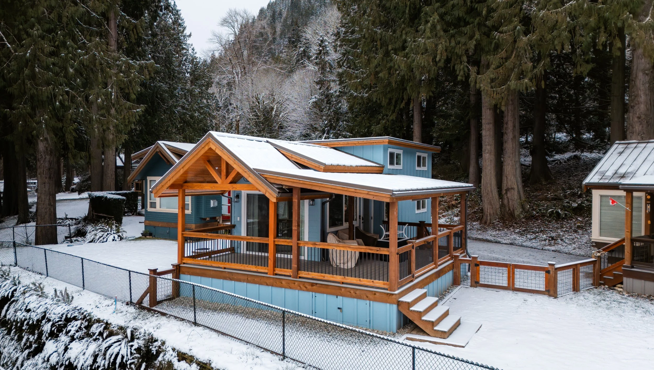 A blue house with a wooden porch and stairs in a snowy forested area. Tall trees surround the house, and a fence encloses the yard.