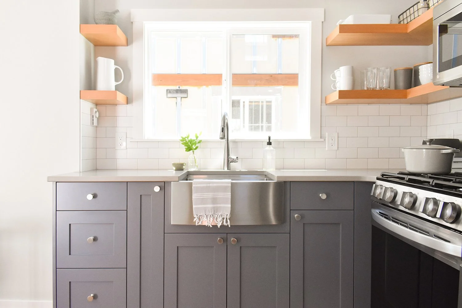 Modern kitchen with gray cabinets, white subway tile backsplash, stainless steel farmhouse sink, open wooden shelves with dishes and kitchenware, window above sink, and gas stove with a pot.