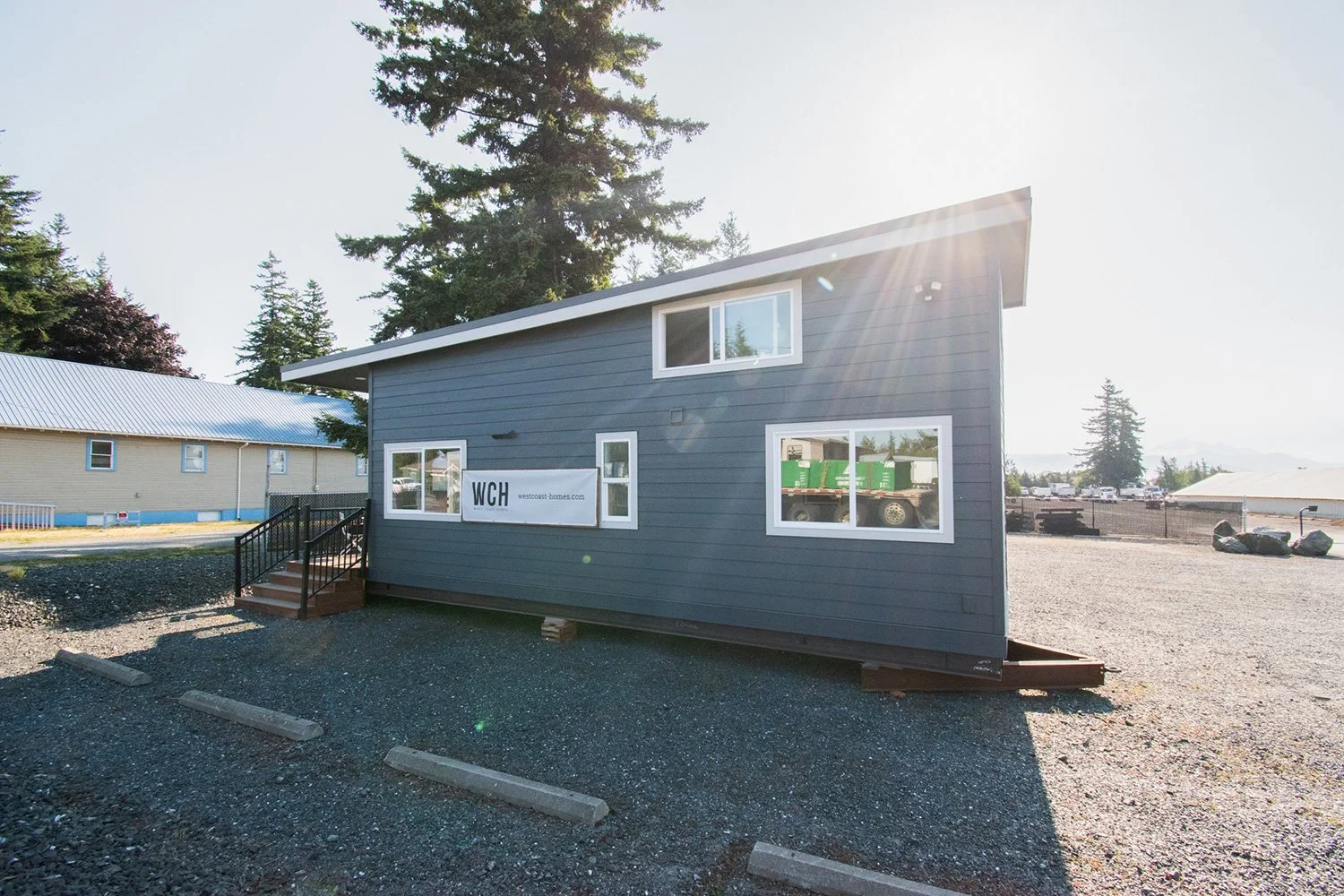 New gray house under construction with white window frames, a small staircase, and a sign that reads "WCH" on an unfinished lot with gravel ground and pine trees in the background.