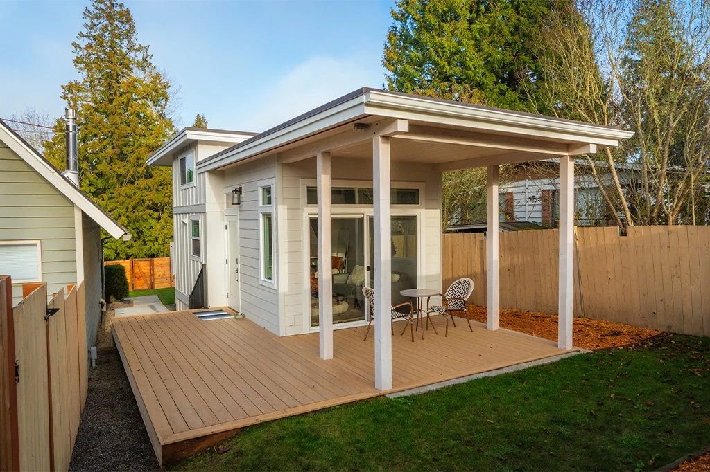 A modern house backyard with a wooden deck and outdoor seating, surrounded by a wooden fence and trees.