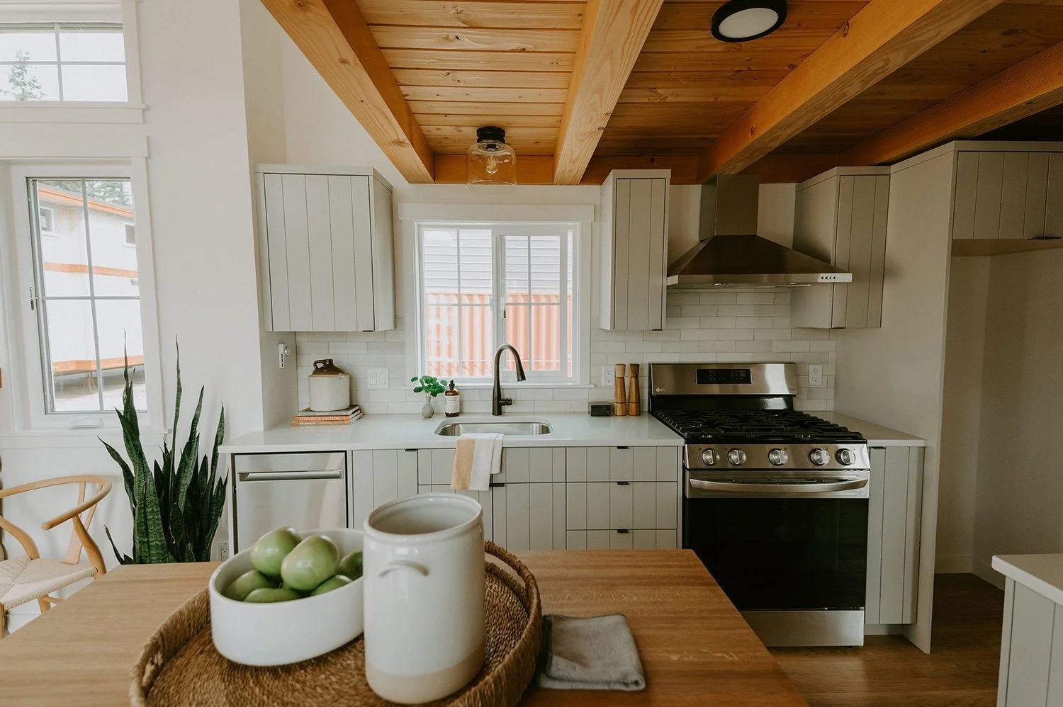 Modern kitchen with white cabinets, stainless steel stove, wooden countertop, and natural wood ceiling beams. There is a window above the sink, a potted plant, and a bowl of green apples on the dining table.