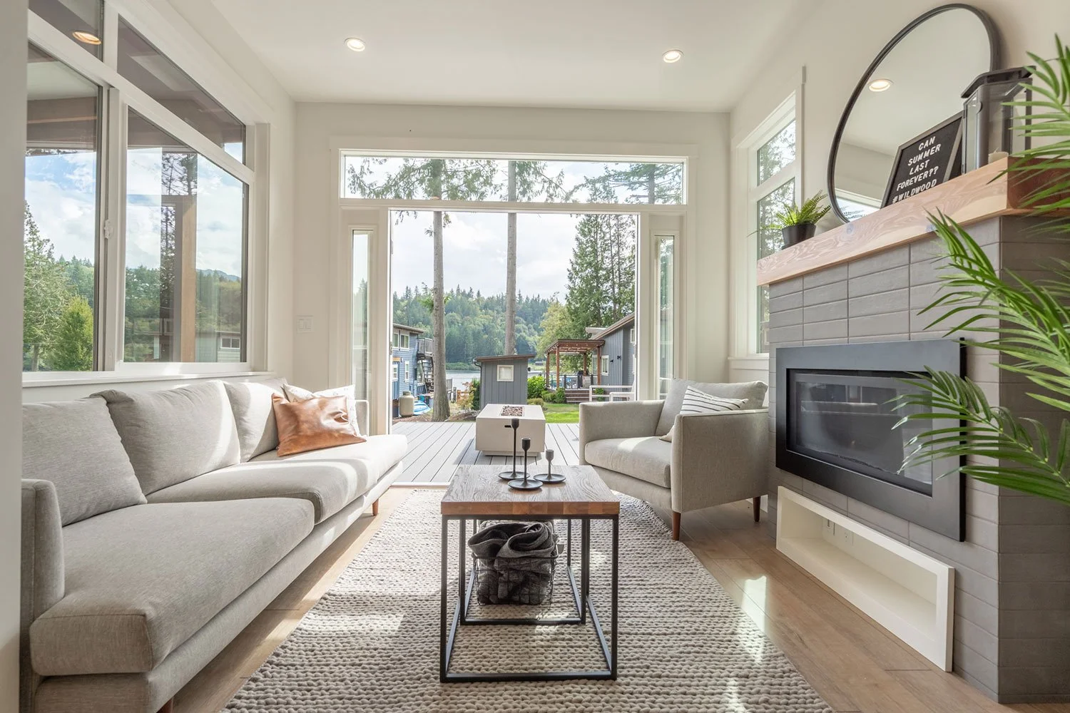 Bright living room with large windows and a sliding glass door opening to a deck and yard with trees and water in the background. Contains a light-colored sofa with a brown pillow, a matching armchair, a wooden coffee table, a fireplace with a mirror and decorations on the mantel, and a potted plant.