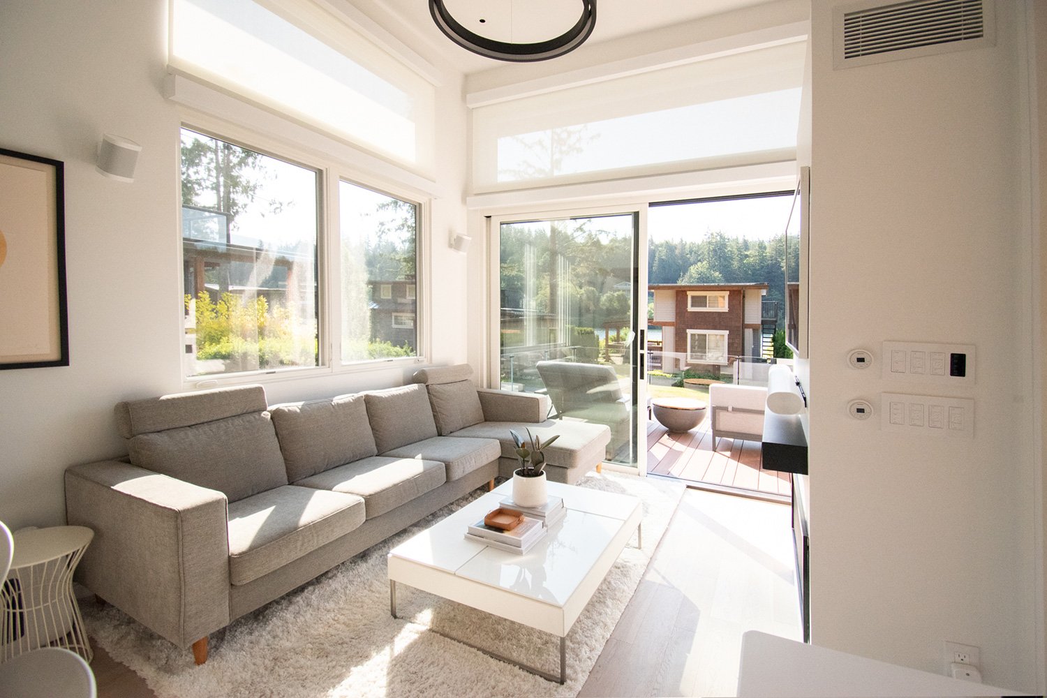 Bright living room with large windows and sliding glass door leading to a balcony with outdoor furniture, beige sofa, coffee table with books and plant, and a white rug.