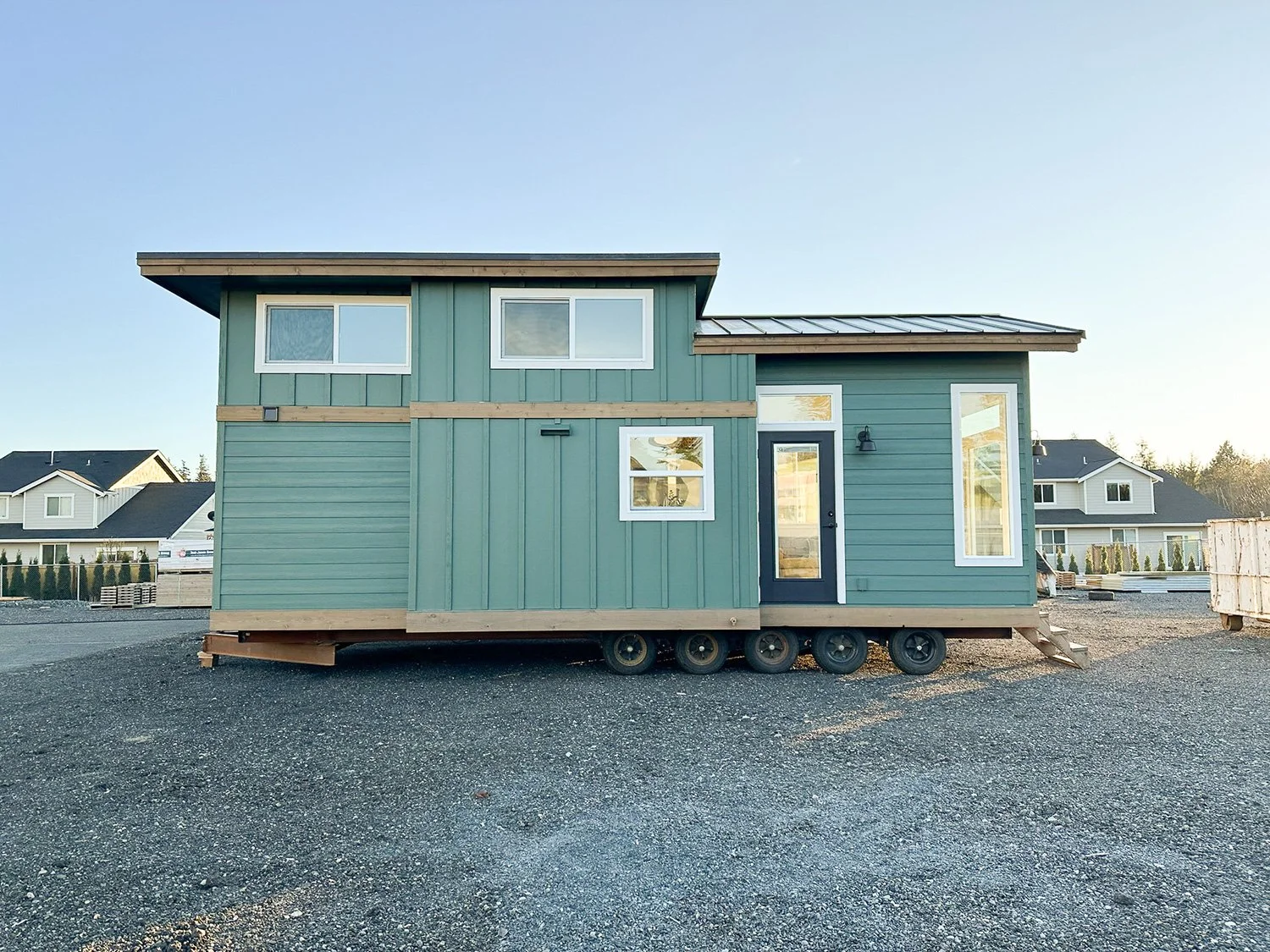 A tiny modern house on wheels painted in green, set on a gravel lot with neighboring houses visible in the background, under a clear sky.