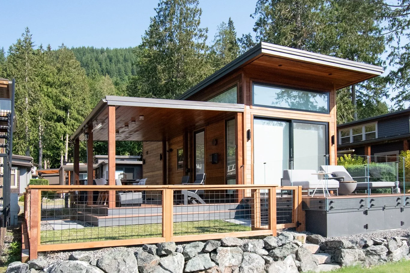 Modern wooden house with large glass windows and a spacious outdoor deck, surrounded by trees and mountains.