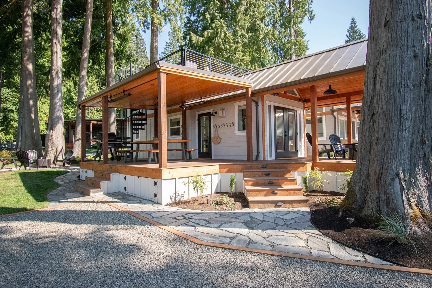 A modern house with a large wooden deck in a forested area with tall trees, outdoor seating and a stone pathway.