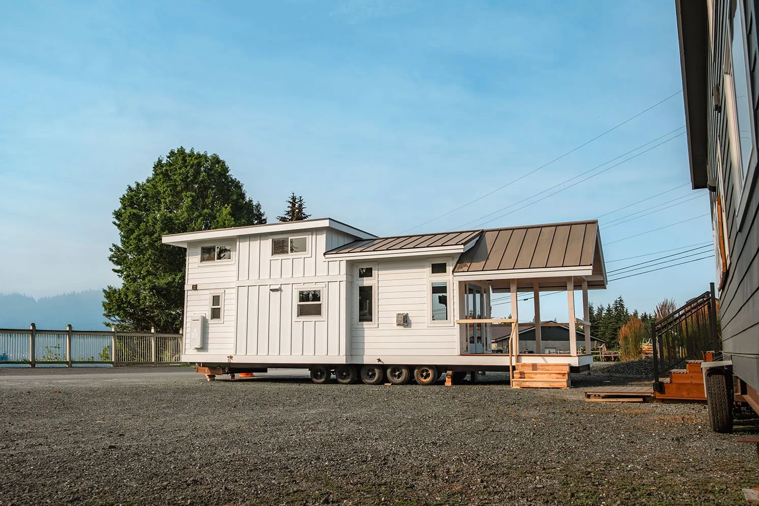 White tiny house on wheels with a raised front porch, set on a gravel lot with a fence and trees in the background.