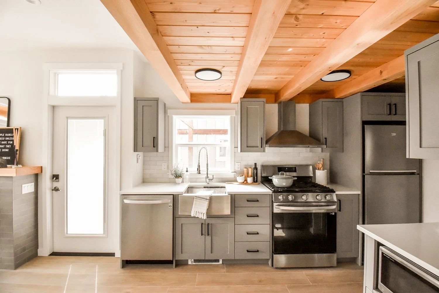 Modern kitchen with gray cabinets, stainless steel appliances, wooden ceiling beams, white countertops, and a window above the sink.
