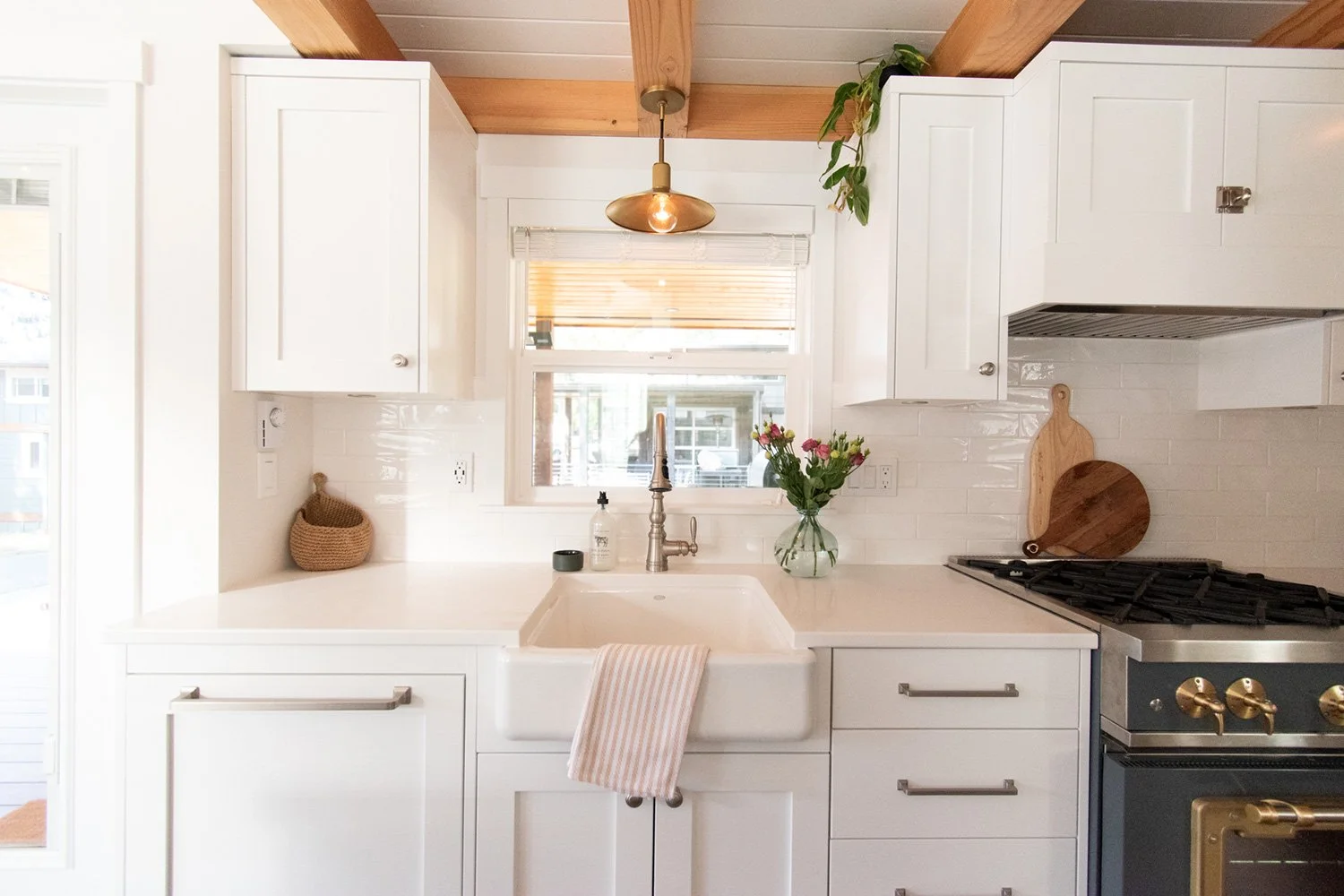 A white kitchen with a farmhouse sink, a window above it, a vase with pink and white flowers, and a stove with golden knobs.