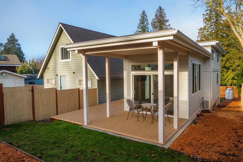 Backyard view of a two-story house with a new wooden deck and patio furniture, surrounded by a wooden fence, with trees in the background.