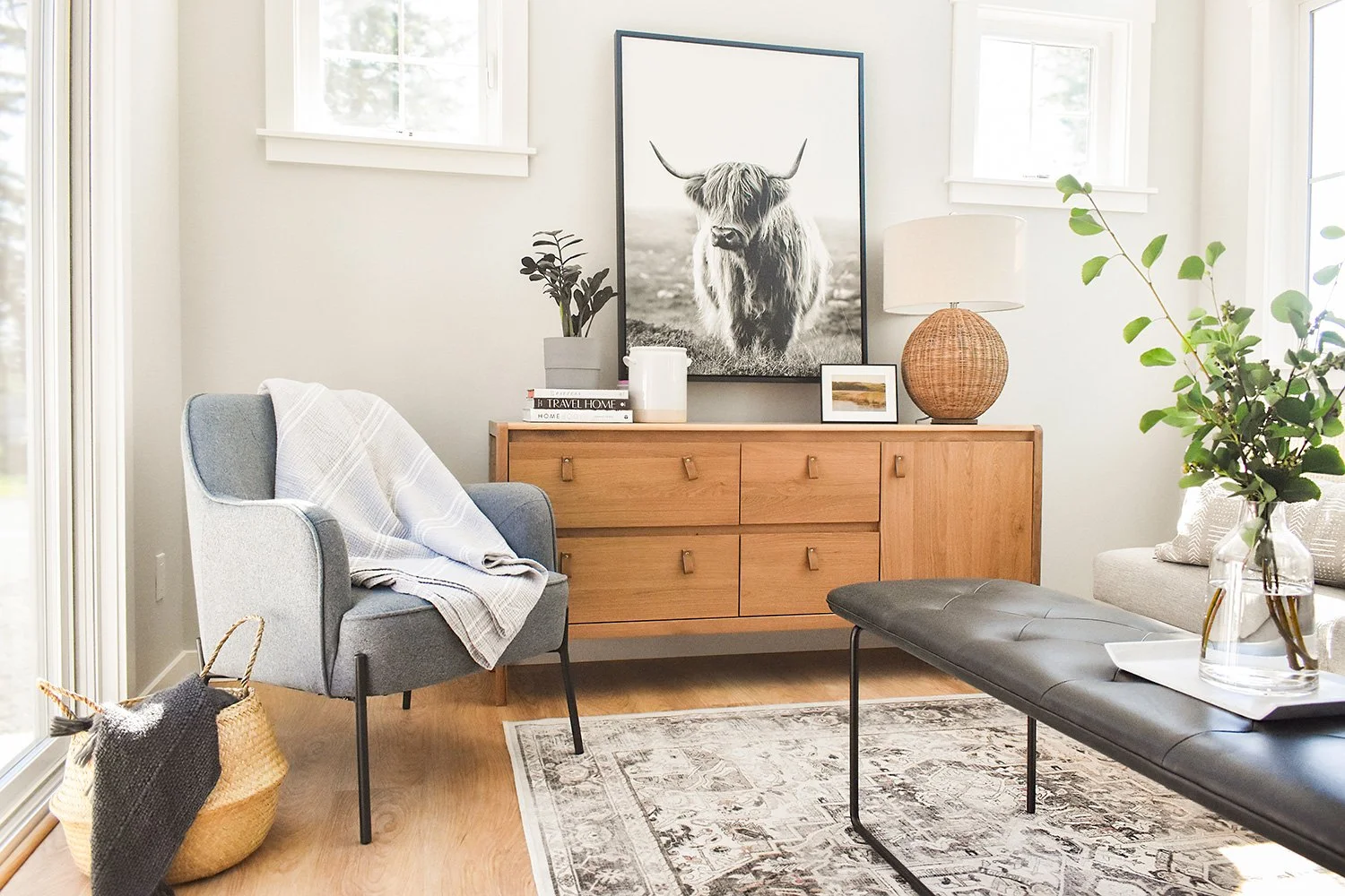 A modern living room with light-colored walls, a wooden sideboard, a large black and white Highland cow portrait, a gray armchair with a blanket, a woven basket, a glass vase with large greenery, a black leather bench, and a patterned area rug.