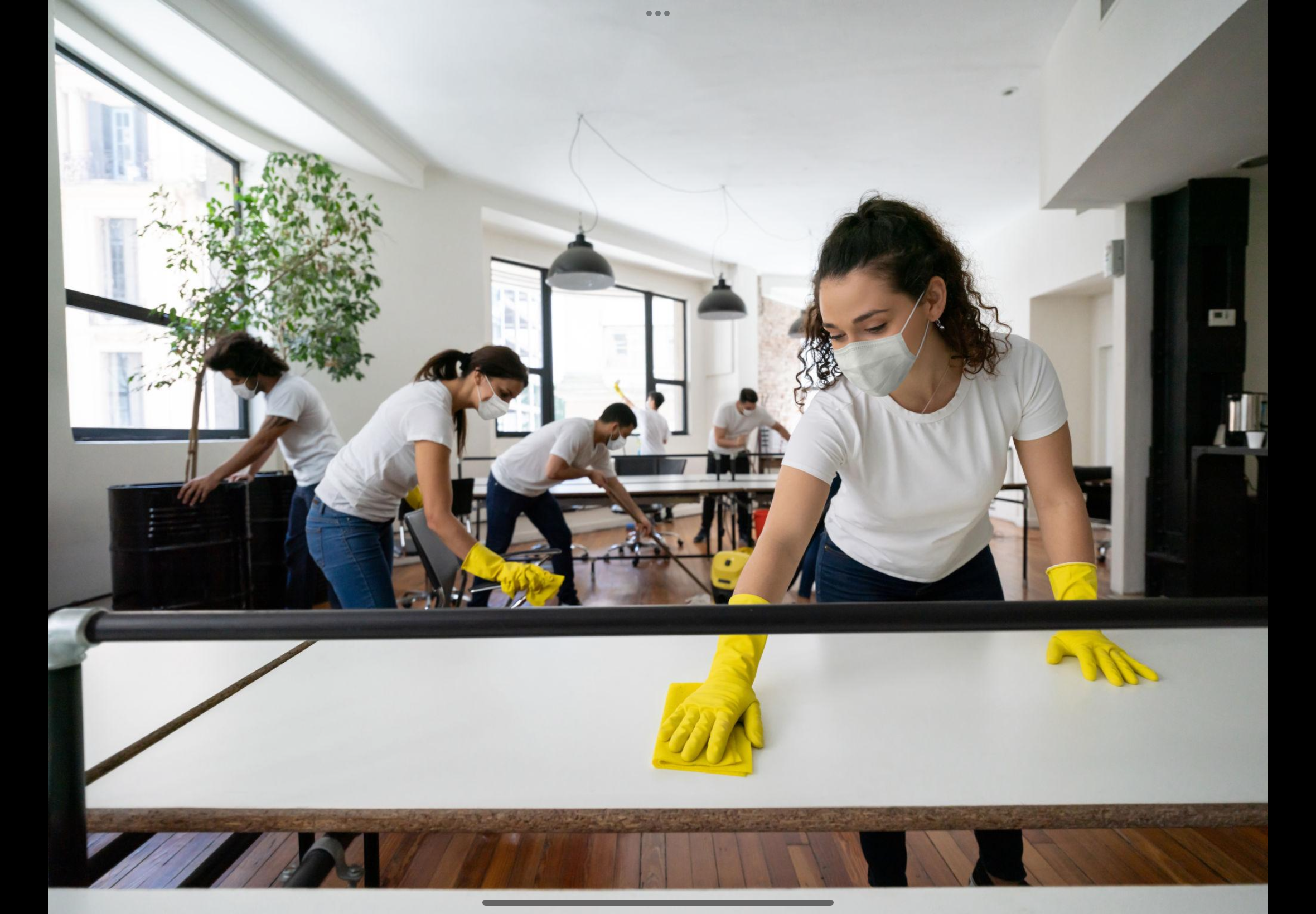 People wearing white shirts, masks, and yellow gloves cleaning or working on a table in a bright room with large windows and hanging lights.