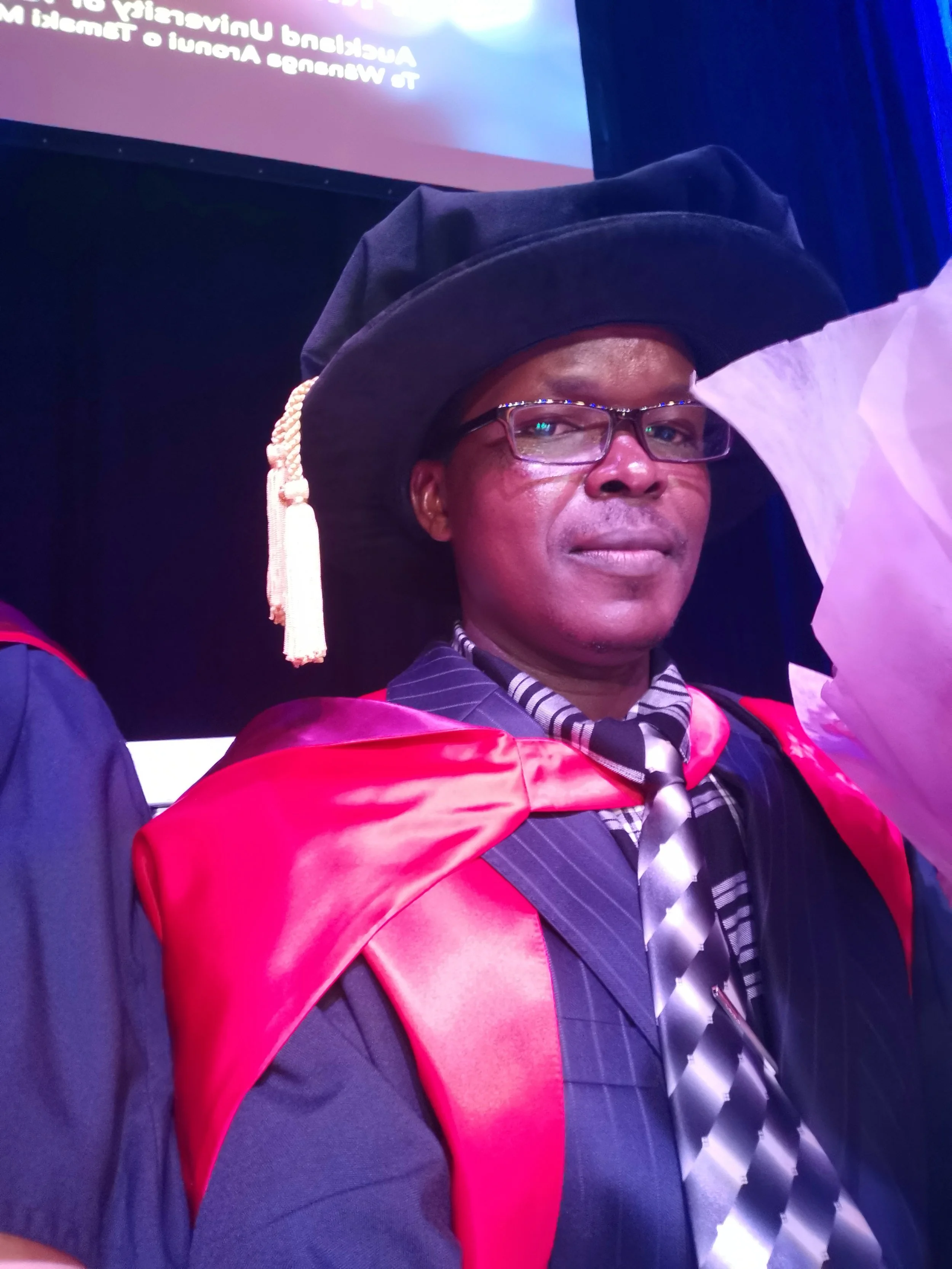 Person wearing academic regalia, including a black mortarboard with a tassel, glasses, a striped shirt, and a suit with a tie, at a graduation ceremony.