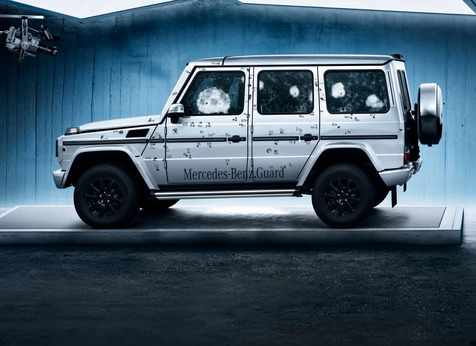 A Mercedes-Benz Guard vehicle with a silver exterior featuring multiple bullet holes and shattered windows, parked on a platform in a dimly lit room with a blue wall and a security camera in the upper left corner.