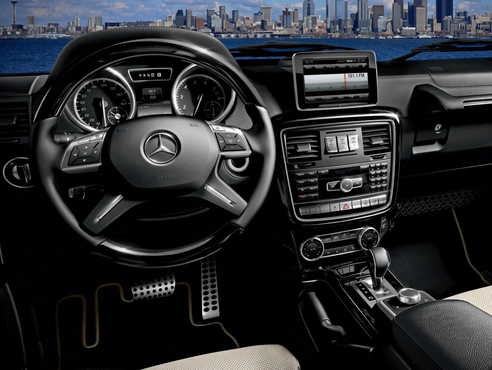 Interior of a luxury Mercedes-Benz vehicle showing a steering wheel, dashboard, infotainment screen, climate controls, and gear shift, with a city skyline visible through the windshield.