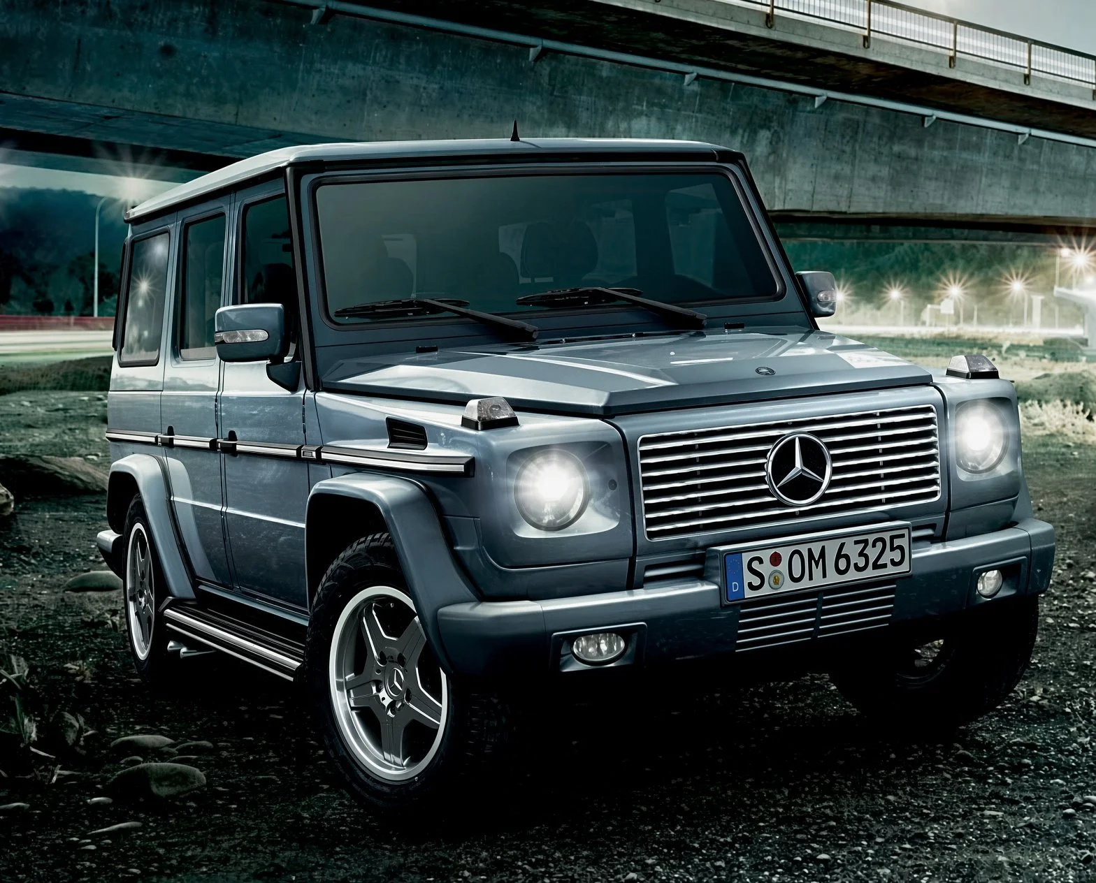 A silver Mercedes-Benz G-Class SUV parked outdoors near a bridge at dusk, with illuminated streetlights in the background.