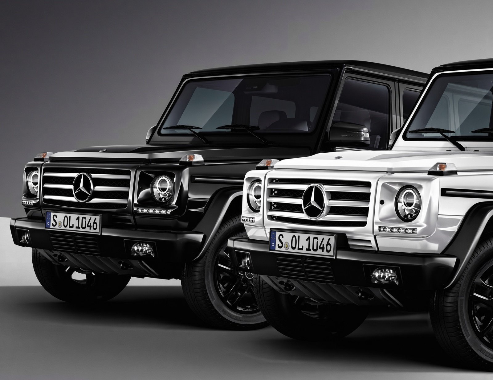 Two Mercedes-Benz G-Class vehicles, one black and one white, parked side by side against a plain gray background.