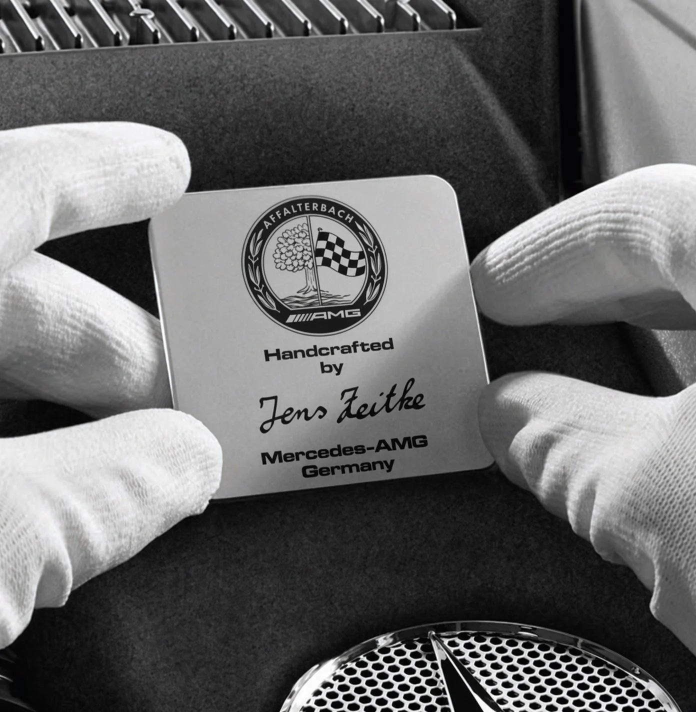 Close-up of a person wearing white gloves holding a certificate or badge that features a logo with a tree and a checkered flag, and text indicating it is handcrafted by Jens Zeitze for Mercedes-AMG Germany.