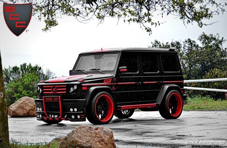 A black Mercedes-Benz G-Class with custom red accents, red wheels, and a matte black finish, parked outdoors near rocks and trees.