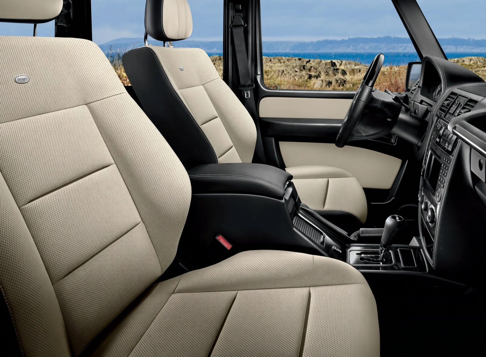 Interior of a vehicle with beige perforated fabric seats, black center console, and dashboard with controls, overlooking an outdoor landscape with rocks, grass, and water in the distance.