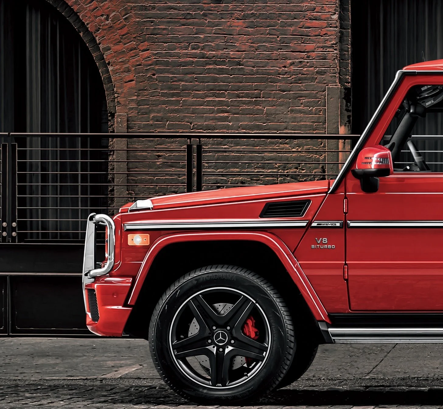 Close-up of the front side of a red Mercedes-Benz G-Class SUV parked against a brick wall with black curtains, showing the wheel, side mirror, and various badging including AMG and V8 Biturbo.