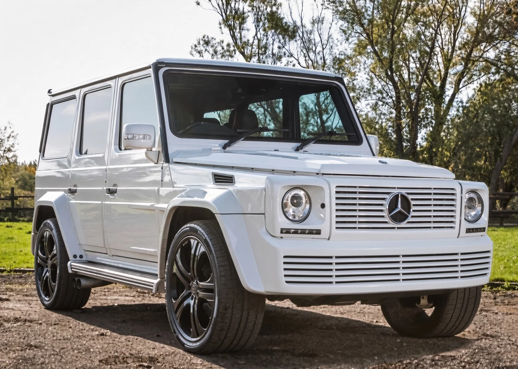 A white Mercedes-Benz G-Class SUV parked outdoors on dirt with trees in the background and a partly cloudy sky.