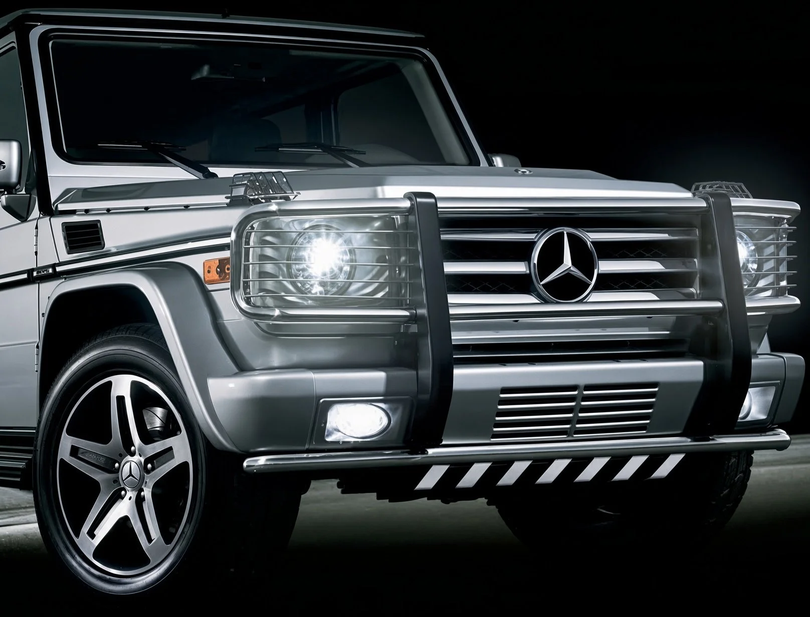 Close-up of a silver Mercedes-Benz G-Class SUV with a custom front grille, bumper guard, and black trim, set against a dark background.