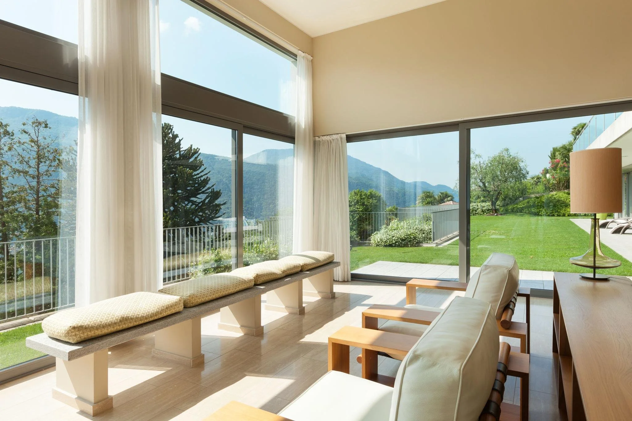 A bright living room with large floor-to-ceiling windows showing a green outdoor yard with trees and mountains in the background. There is a built-in bench with cushions beneath the windows, and a wooden dining table with chairs near the windows. A floor lamp stands beside the table.
