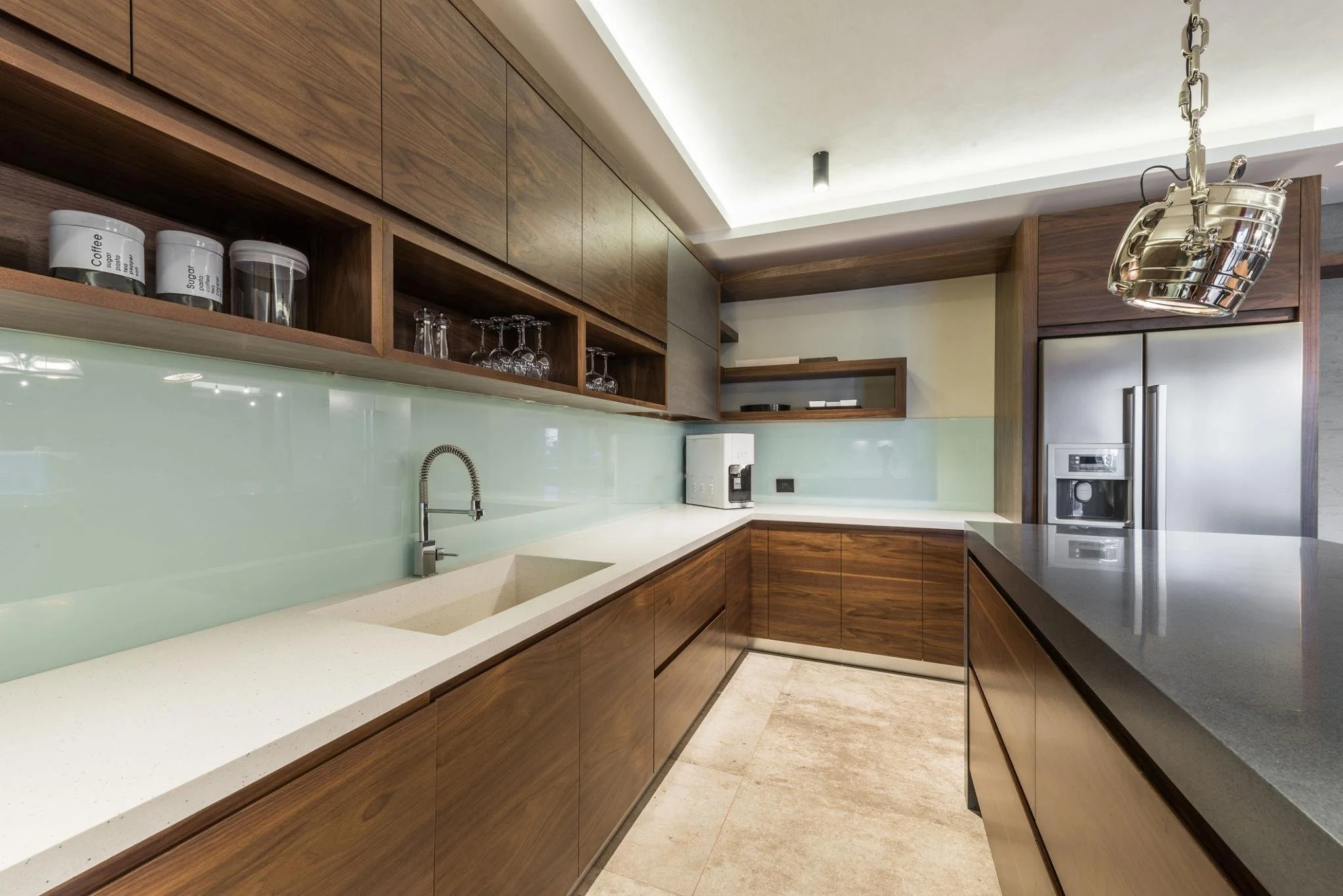 Modern kitchen with wooden cabinets, a white countertop, and a frosted glass backsplash. Includes a sink with a flexible faucet, wine glasses, and kitchen appliances such as a refrigerator and microwave.