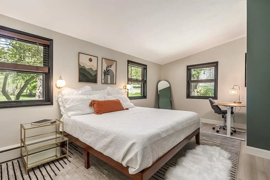 A bright bedroom with a large bed, white bedding, and an orange pillow, with three framed art pieces above the bed, two windows with blinds, a small gold side table with a book, a standing mirror, a desk with a lamp and chair, a rug, and a fluffy white rug on the floor.