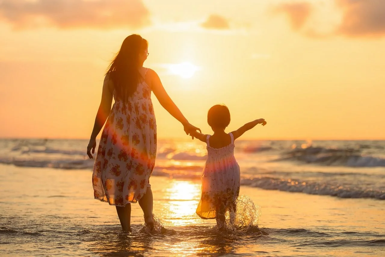 A woman and a young girl walking hand in hand in shallow ocean water at sunset, with the sun Low in the sky and a calm, colorful sky in the background.