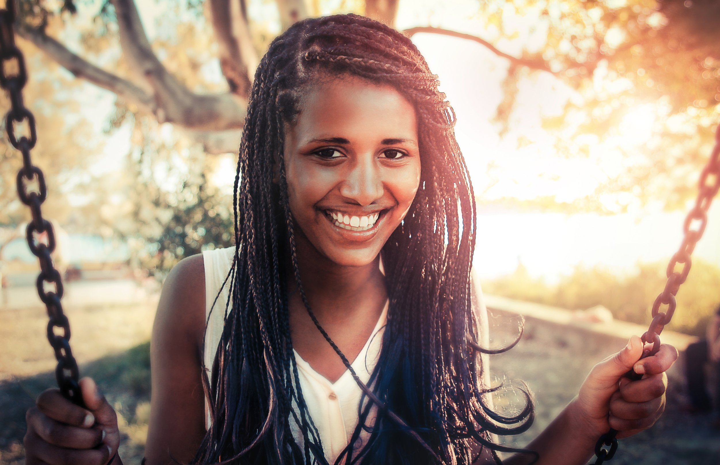 A young woman with braided hair smiling while sitting on a swing in a park during sunset.