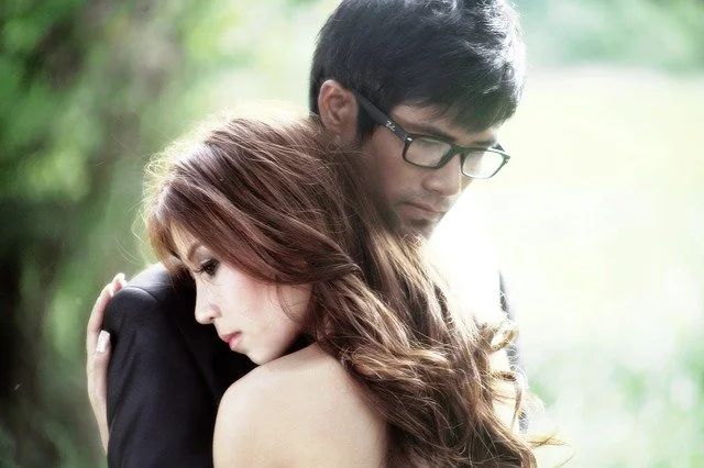 A young couple stands close together outdoors, with the woman resting her head on the man's shoulder.