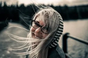 Young woman with glasses and long blonde hair smiling outdoors near a body of water, with a blurred background of trees.