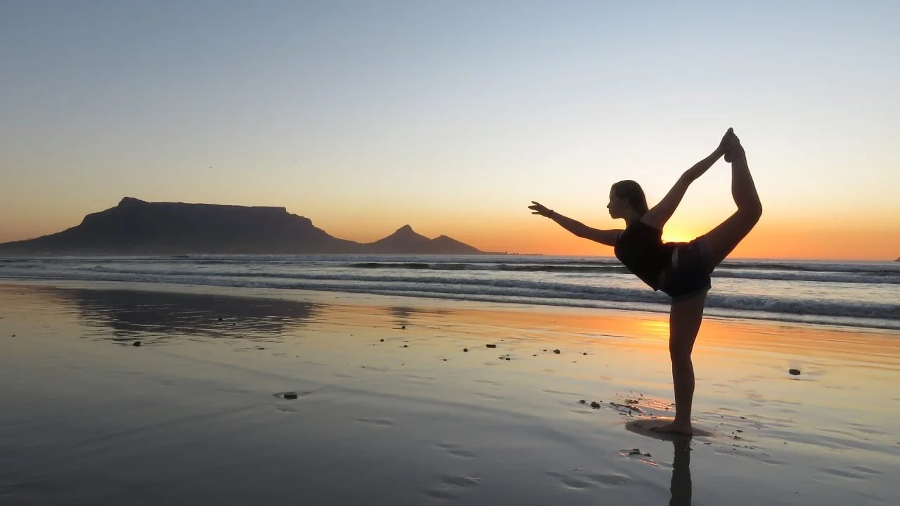 A person practicing yoga on a beach at sunset, performing a backbend pose with one leg raised behind and their arm extended forward, with mountains in the background.