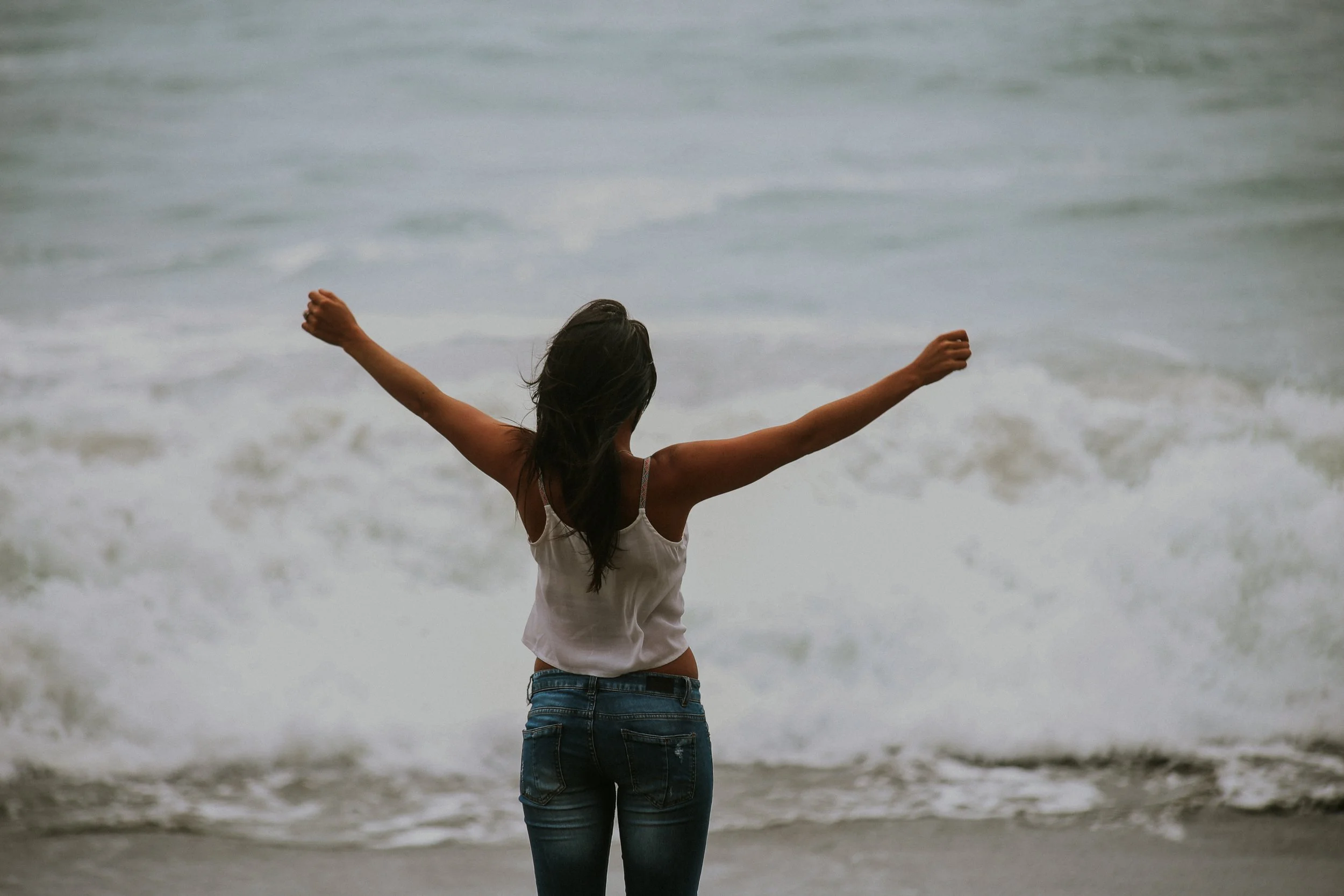 A woman with long dark hair wearing a tank top and jeans standing on the beach with arms raised and open towards the ocean.