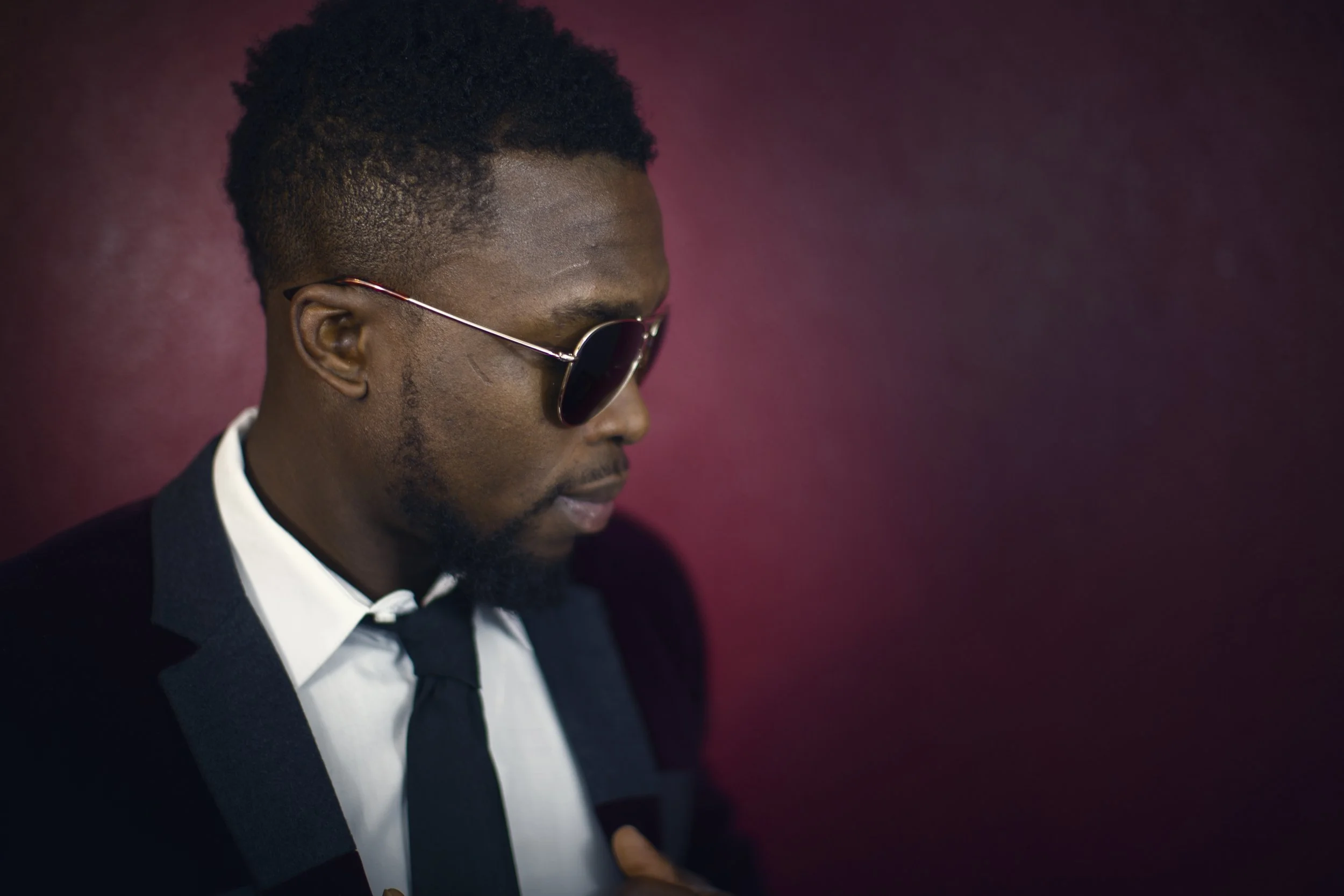 A man wearing sunglasses, a black tuxedo, and a white shirt with a bowtie, posing against a maroon background.