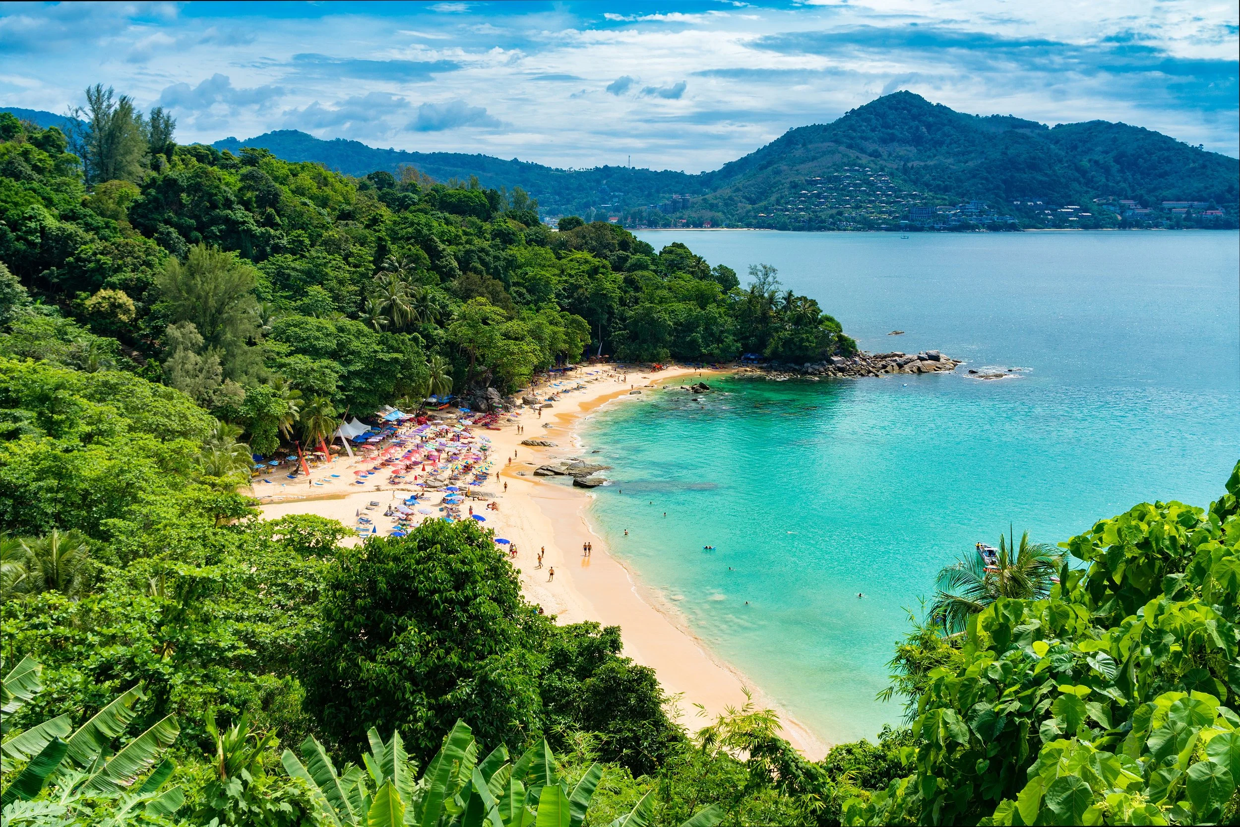 A tropical beach with clear turquoise water, white sand, and colorful umbrellas. Dense green foliage and trees line the beach, with hills and mountains in the background under a partly cloudy sky.