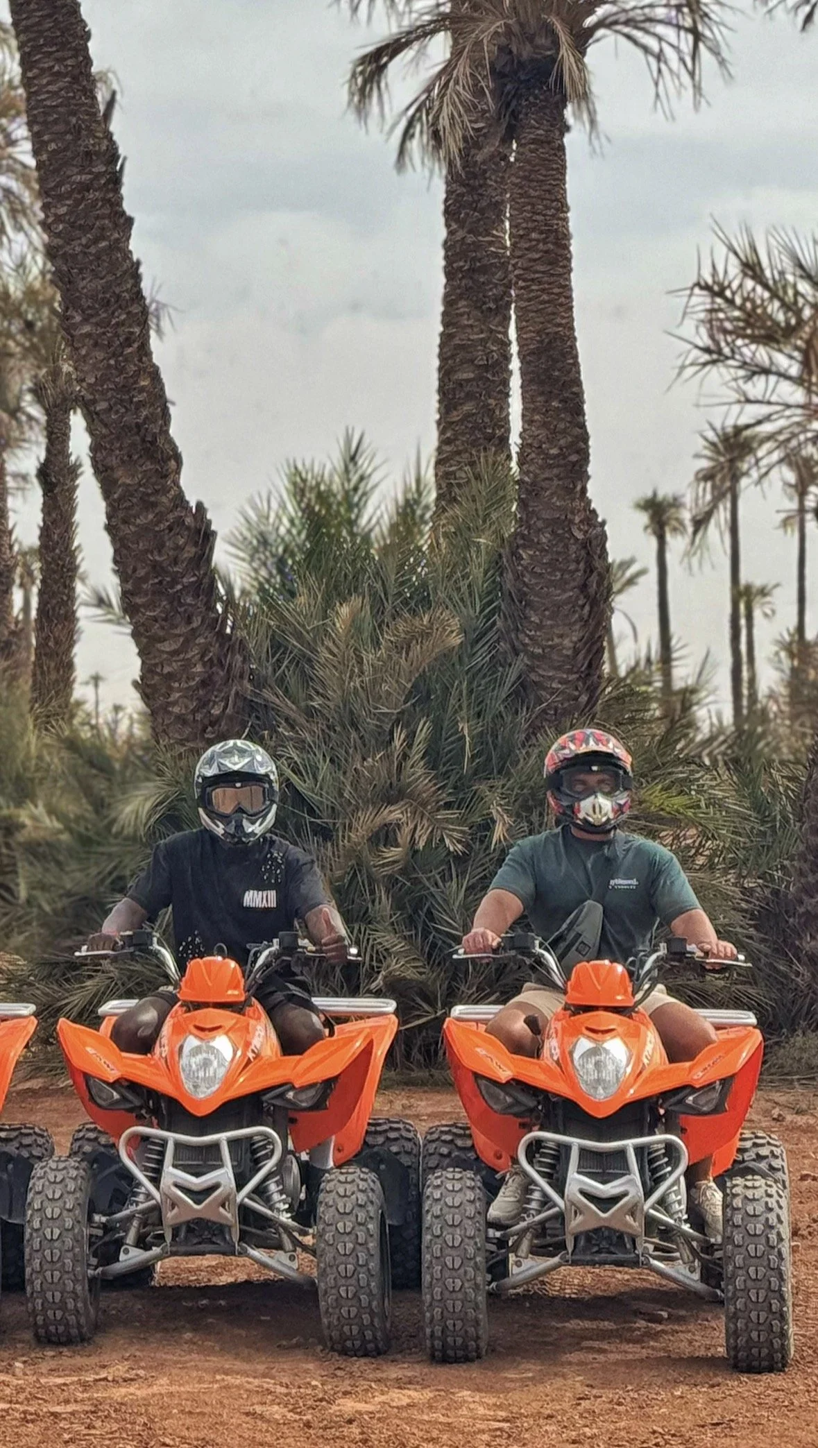 Two people wearing helmets and sunglasses sitting on orange all-terrain vehicles (ATVs) in front of tall palm trees on a dirt ground.